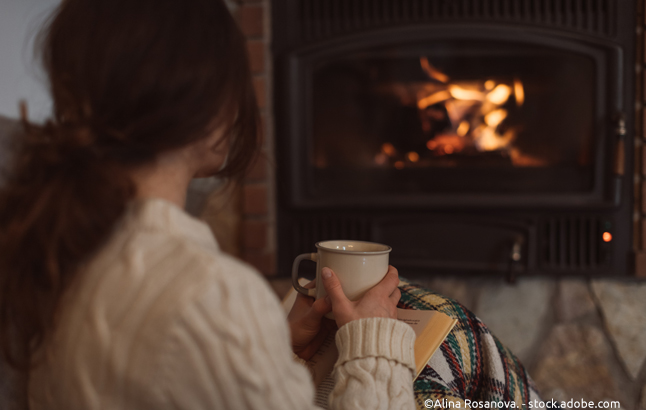 Junge Frau wärmt sich an warmem Becher in Decke gehüllt vor Kamin auf
